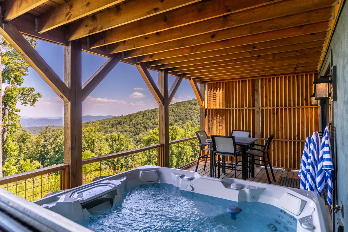 A spacious outdoor deck with a hot tub and dining table, overlooking expansive mountain views. The area is bathed in sunset light, creating a tranquil atmosphere perfect for relaxing or setting up a laptop for work. The surroundings emphasize privacy and serenity, with nature in the backdrop.