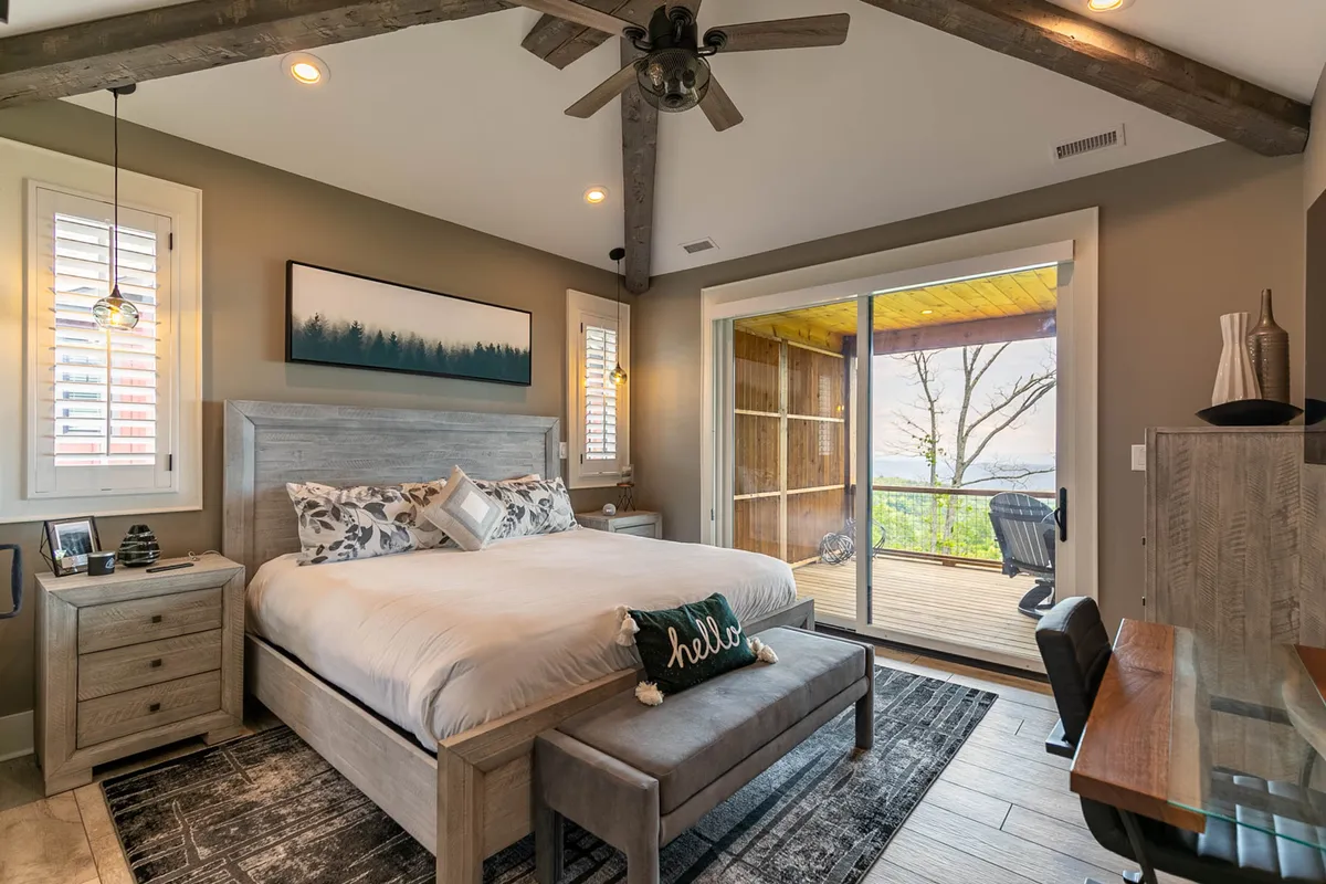 A cozy bedroom featuring a modern desk and chair set up against large glass sliding doors that open to a balcony, providing a serene workspace with natural light and sweeping views. Above the bed, tasteful artwork decorates the space, and the bedding is accented with decorative pillows for a comfortable feel.