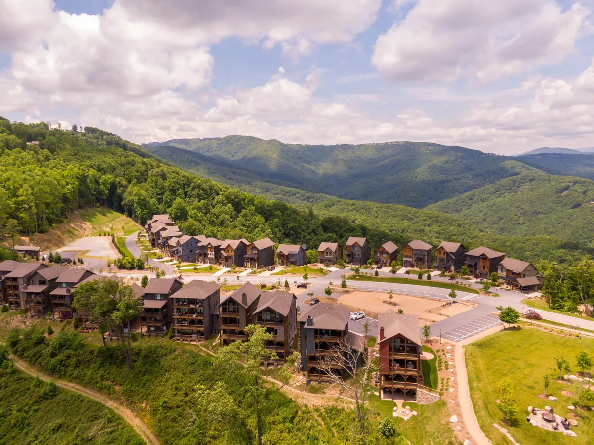A cluster of multi-story wooden cabins surrounded by mountains and lush greenery, with ample space for parking and outdoor activities. The scene suggests immediate access to hiking trails and communal green spaces.
