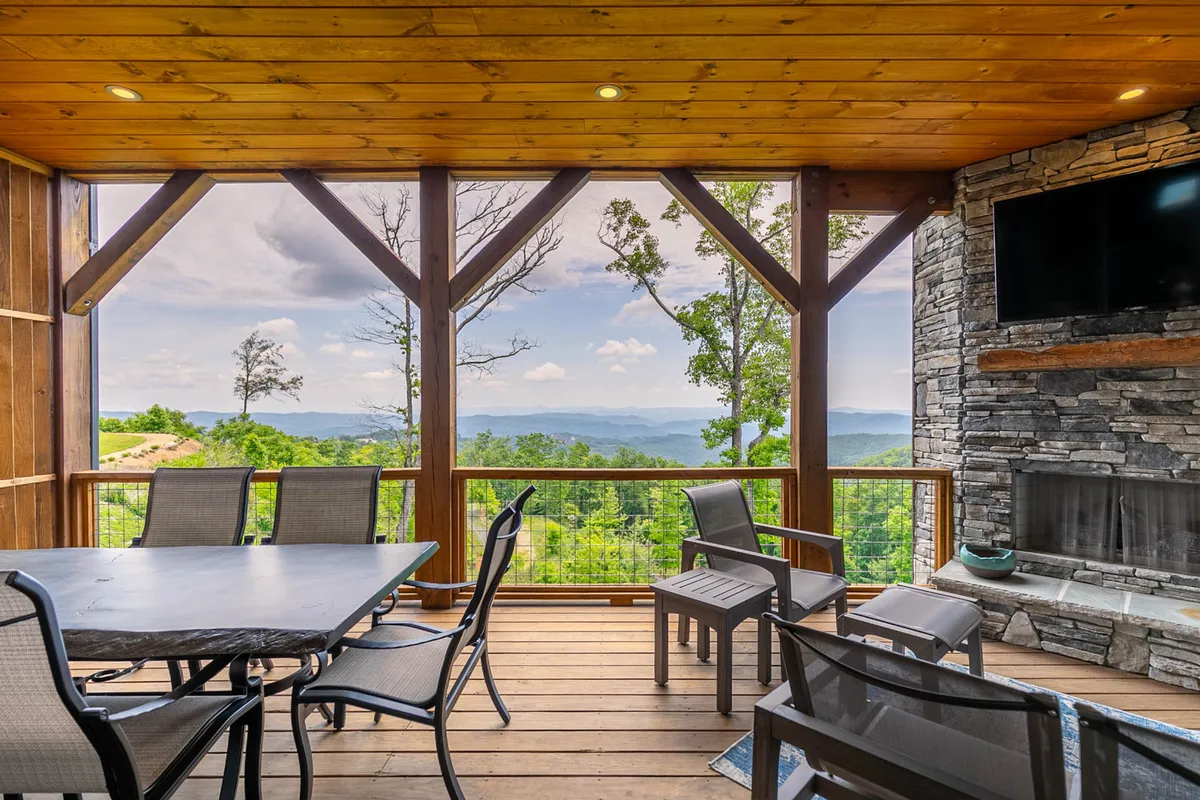 A spacious deck with a dining table set against a backdrop of breathtaking mountain views, accompanied by an outdoor stone fireplace and television. The scene evokes the pleasure of al fresco dining and relaxing in nature’s embrace.