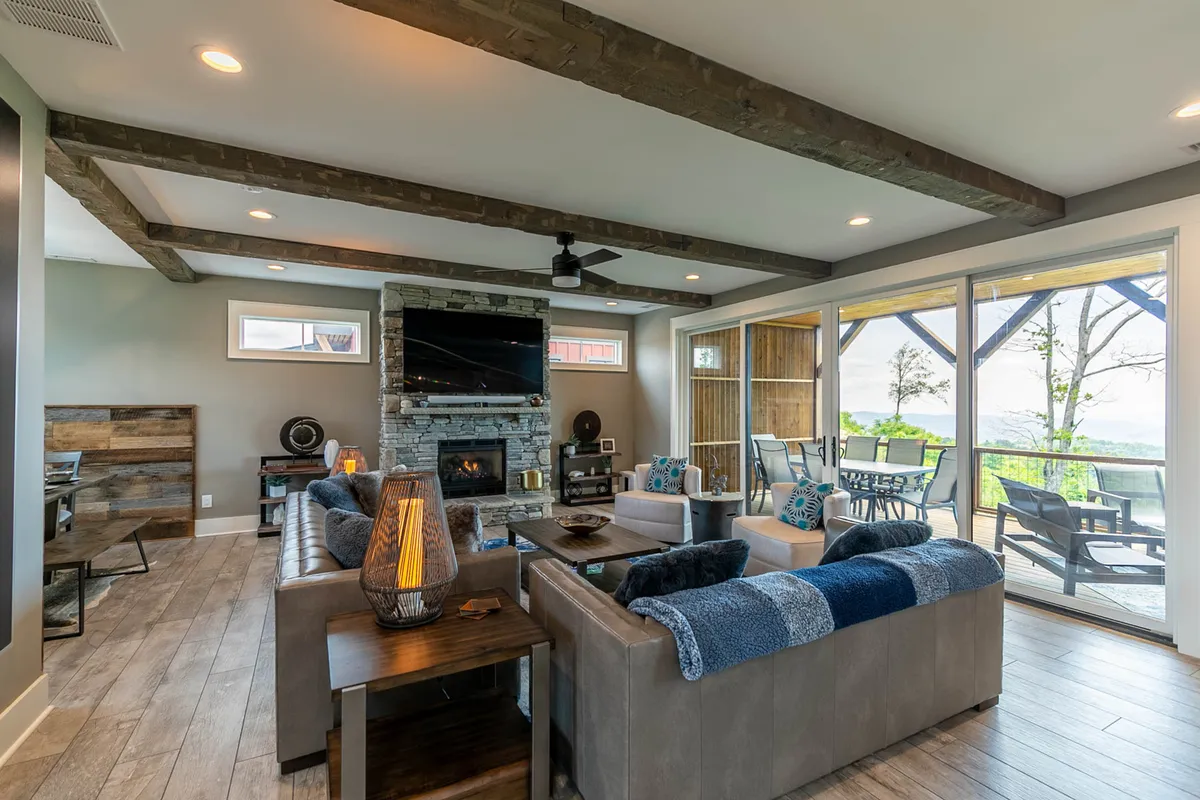 A spacious living room anchored by a dramatic stone fireplace, featuring plush seating arranged for group gatherings, exposed wooden beams overhead, and expansive windows revealing breathtaking mountain views. Sliding glass doors open onto the main deck, blurring the line between indoor comfort and outdoor splendor.