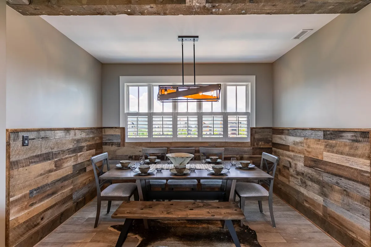 A warm and inviting dining area with a large wooden table set under an elegant chandelier, surrounded by windows letting in natural light and offering glimpses of the mountain landscape. The setting is ideal for family meals or special celebrations.
