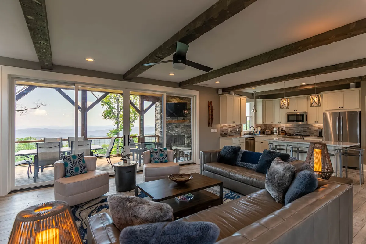 An open-plan great room featuring a comfortable seating area with large windows framing mountain views, a modern kitchen in the background, stylish lighting fixtures, and wooden beams overhead. The room exudes warmth, blending relaxation areas with function and scenery.