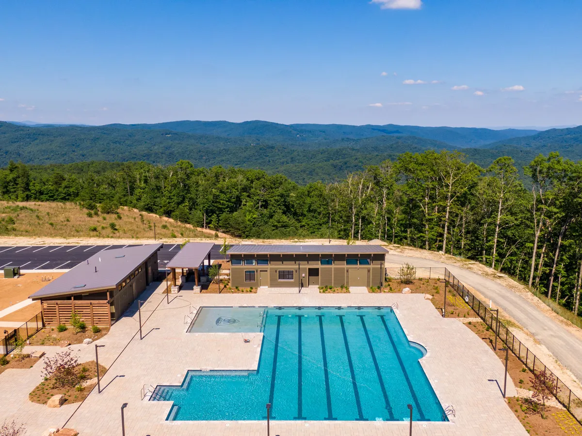 A brand-new outdoor swimming pool is surrounded by a modern building, poolside lounge areas, and sweeping views of nearby mountains. Guests are seen swimming, sunbathing, and gathering with friends, evoking a sense of vibrant social fun and relaxation.