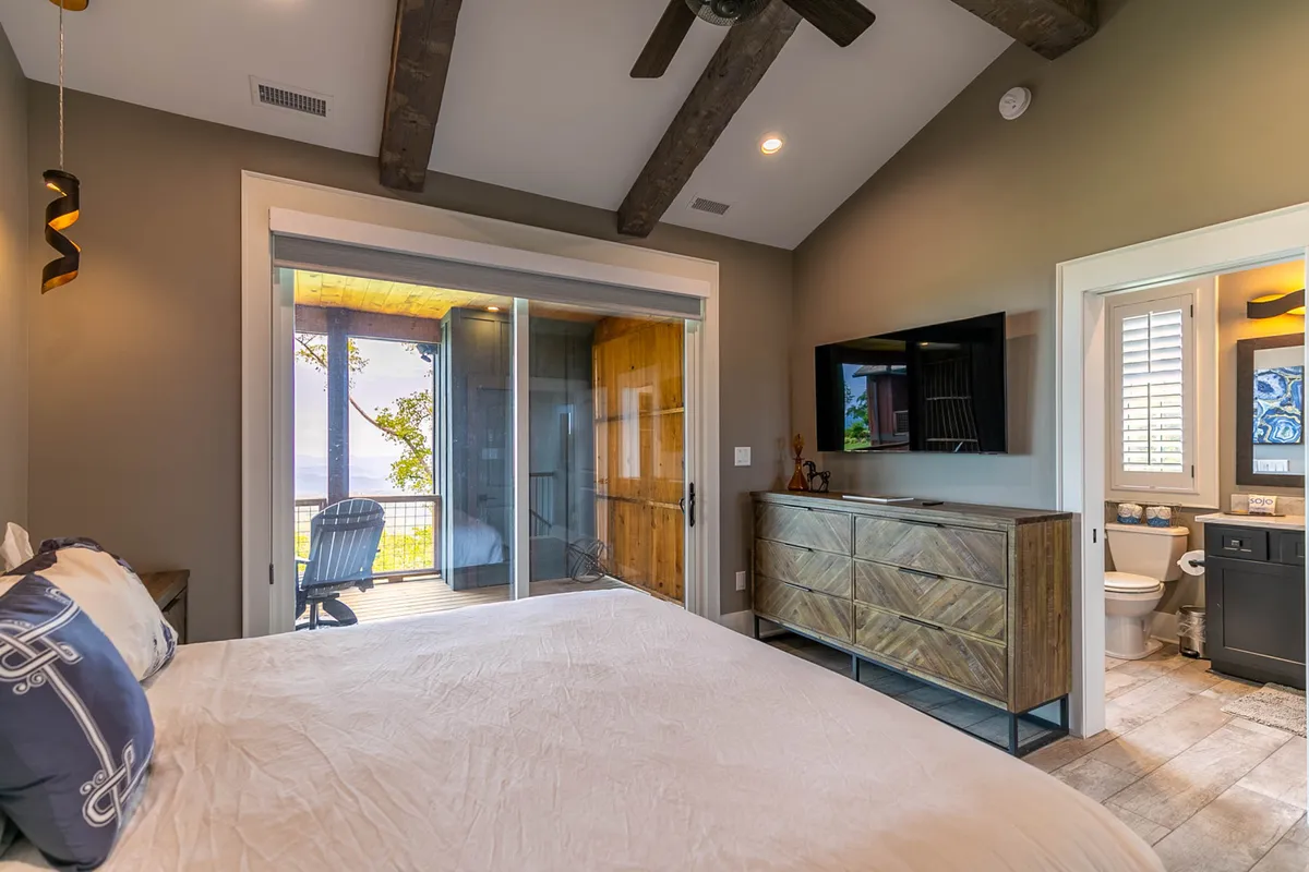 A peaceful master bedroom with a cozy bed, sliding glass doors leading to a private balcony, a wooden dresser, and a television. The space is bathed in natural light, highlighting the room's relaxing and private ambiance.